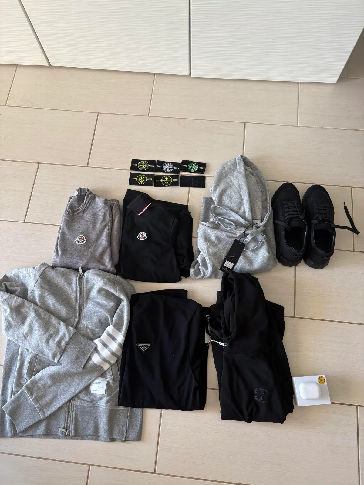 Flat lay of clothing, black shoes, loose badges and an earbud box on pale tile